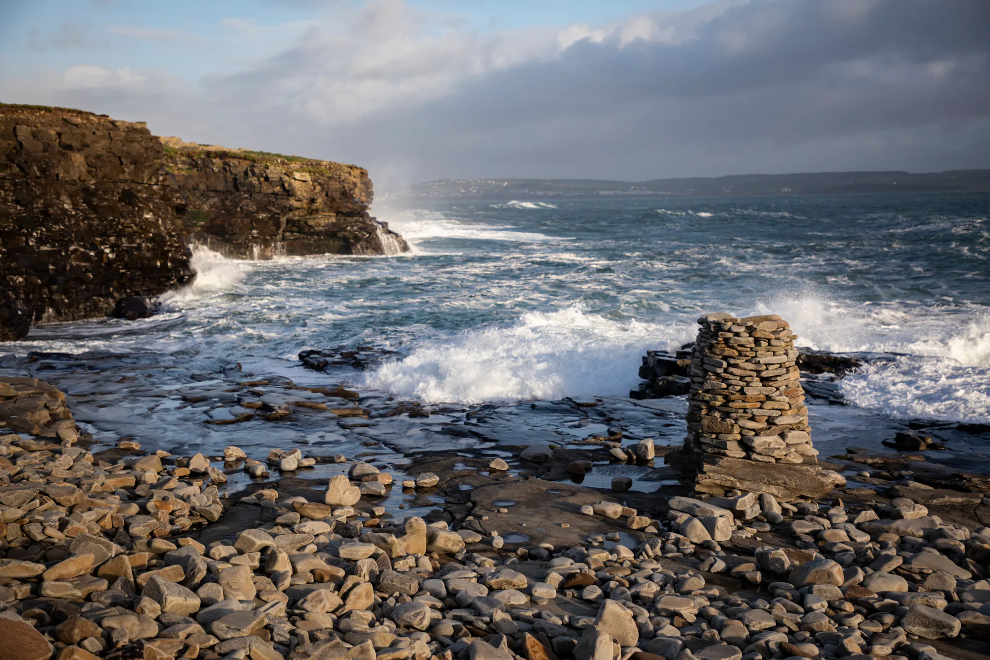 Waves crashing against a pebble beach and cliffs in Ireland with a small rock tower in the foreground.