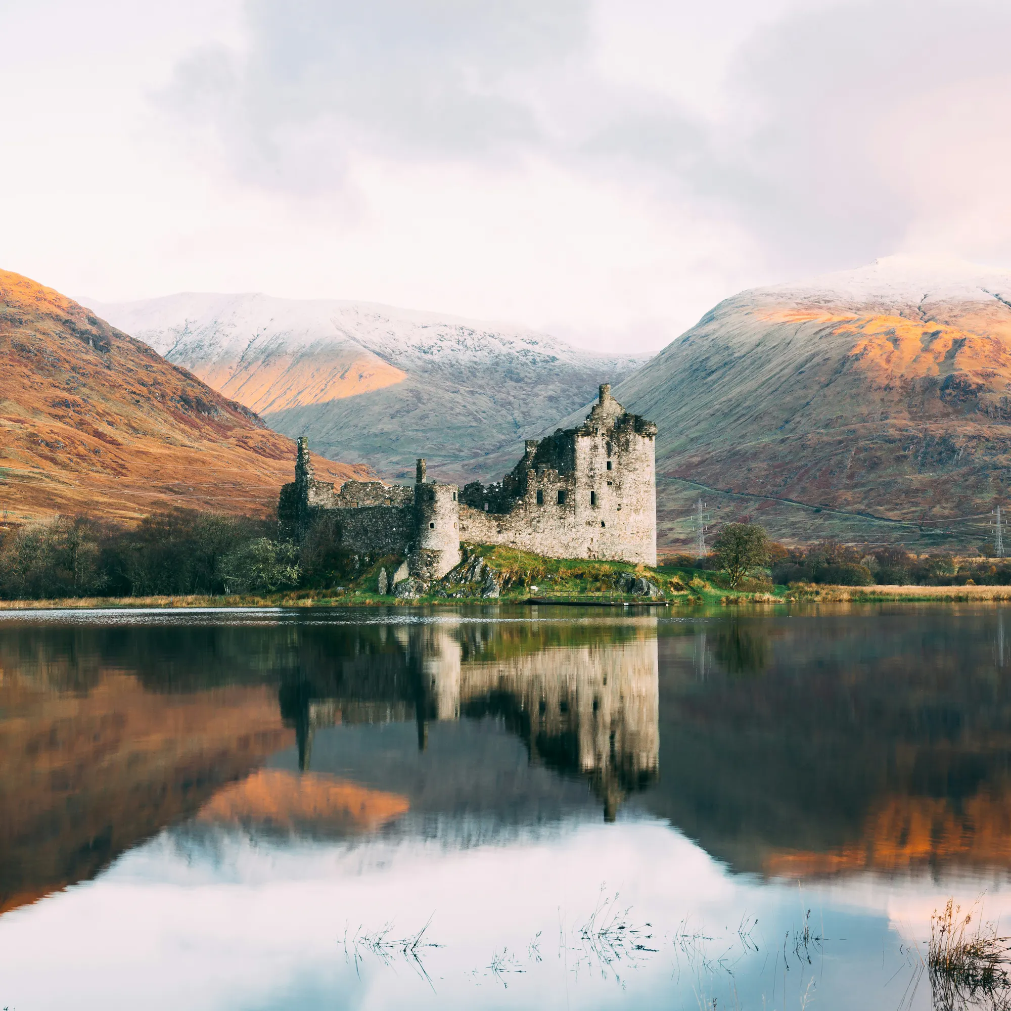 Kilchurn Castle's ruined towers reflected in Loch Awe, framed by autumn hills and snow-capped peaks.