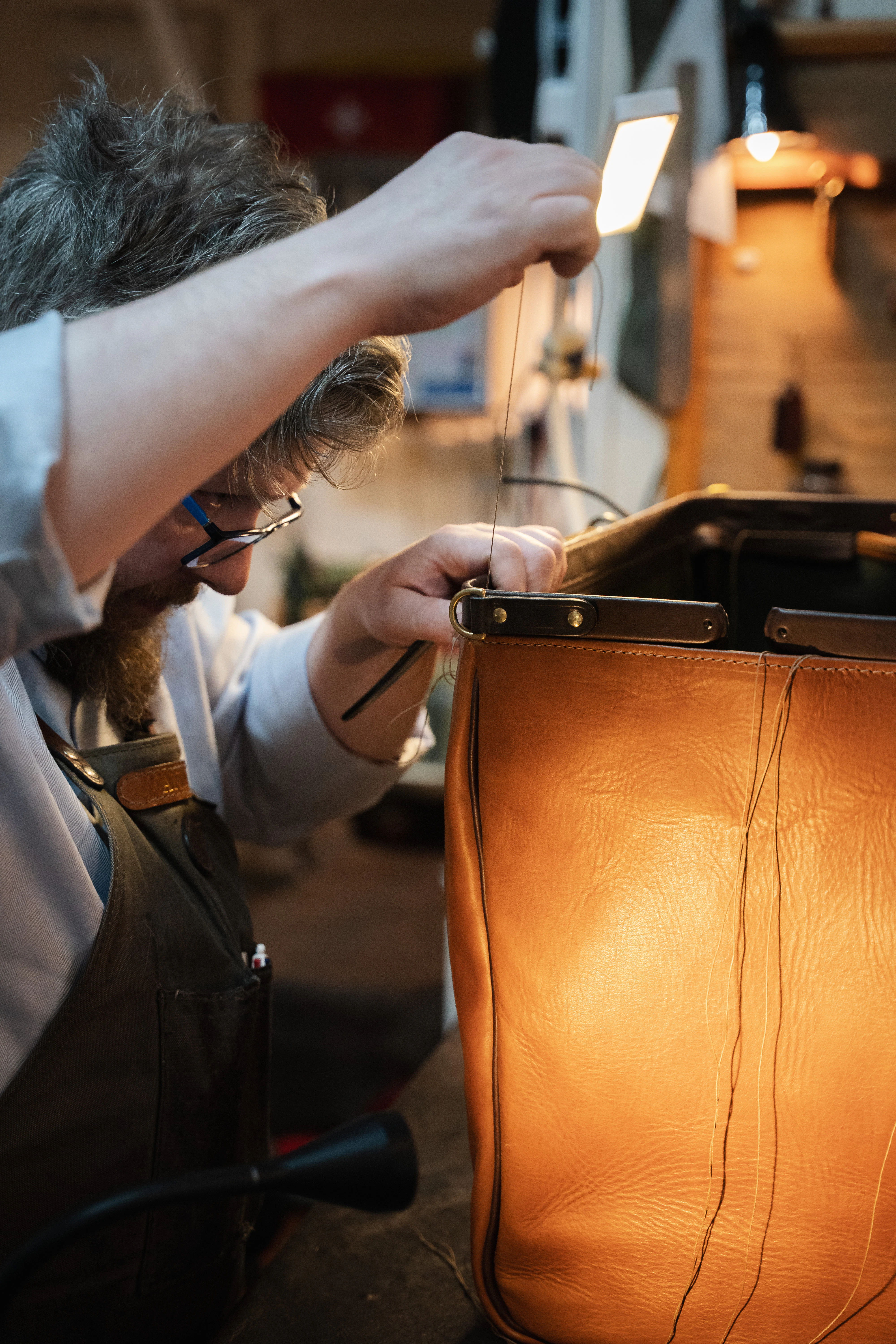 A craftsman hand-stitches a tan leather bag with waxed thread in his workshop.