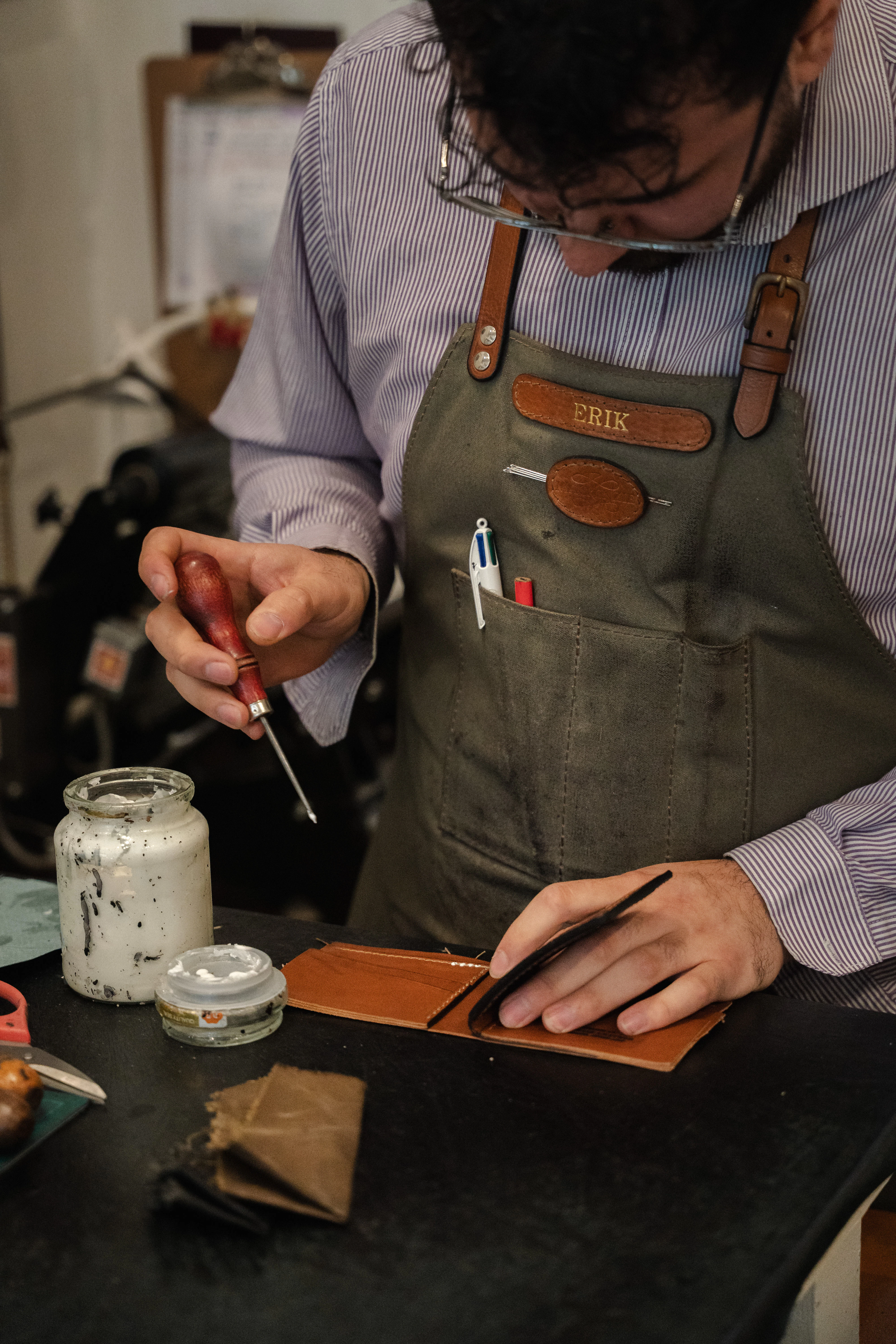 A craftsman in a canvas apron works an awl over a tan leather wallet at his workbench.